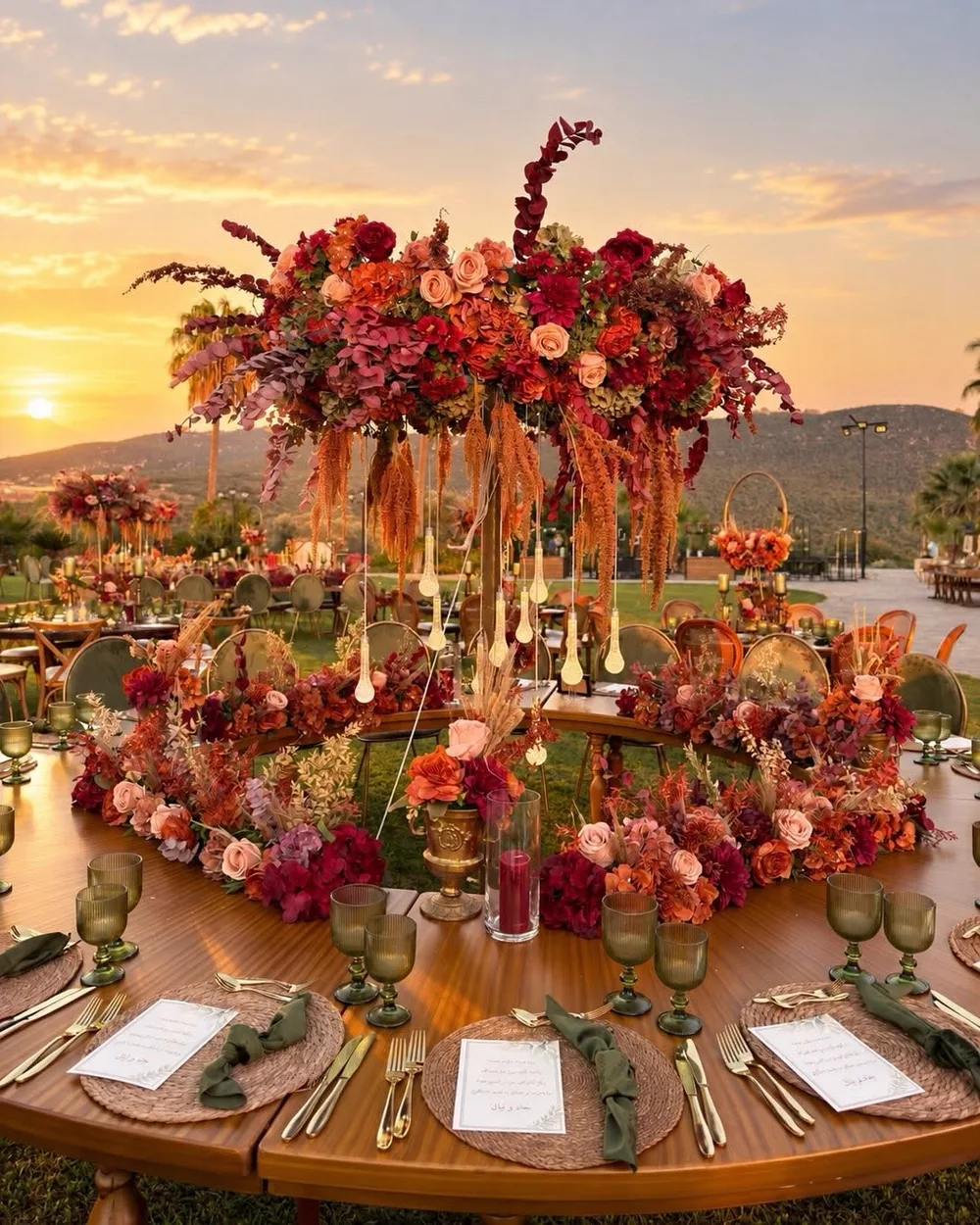 A round wedding table at sunset, set with green glasses and gold cutlery under a tall centrepiece of red, orange and blush roses
